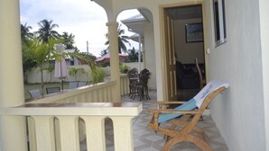 A covered outdoor area with a wooden chair and a table, a view of the backyard with palm trees, and a door leading to another room. - My Ozi Perl Self Catering Guest House (Praslin Island)