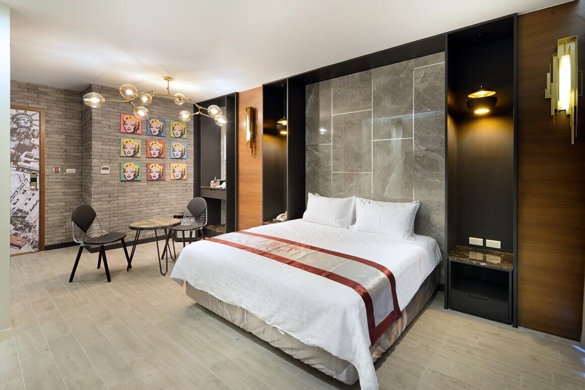A modern hotel room with a large bed, a small table, two chairs, a brick-patterned wall, and artwork.