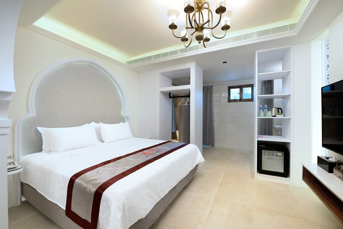 A modern hotel room with a large bed, a chandelier, built-in shelving, and a television.