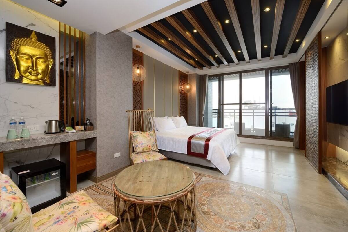 Una habitación de hotel moderna con una cama grande, una zona de estar con una mesa redonda, una pequeña cocina y un ventanal con vistas.
