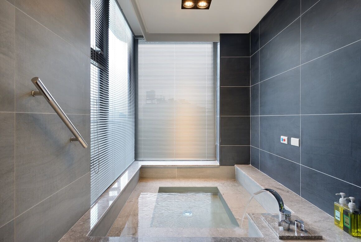 A modern bathroom with a glass-enclosed hot tub, a handrail, and a large window with blinds.