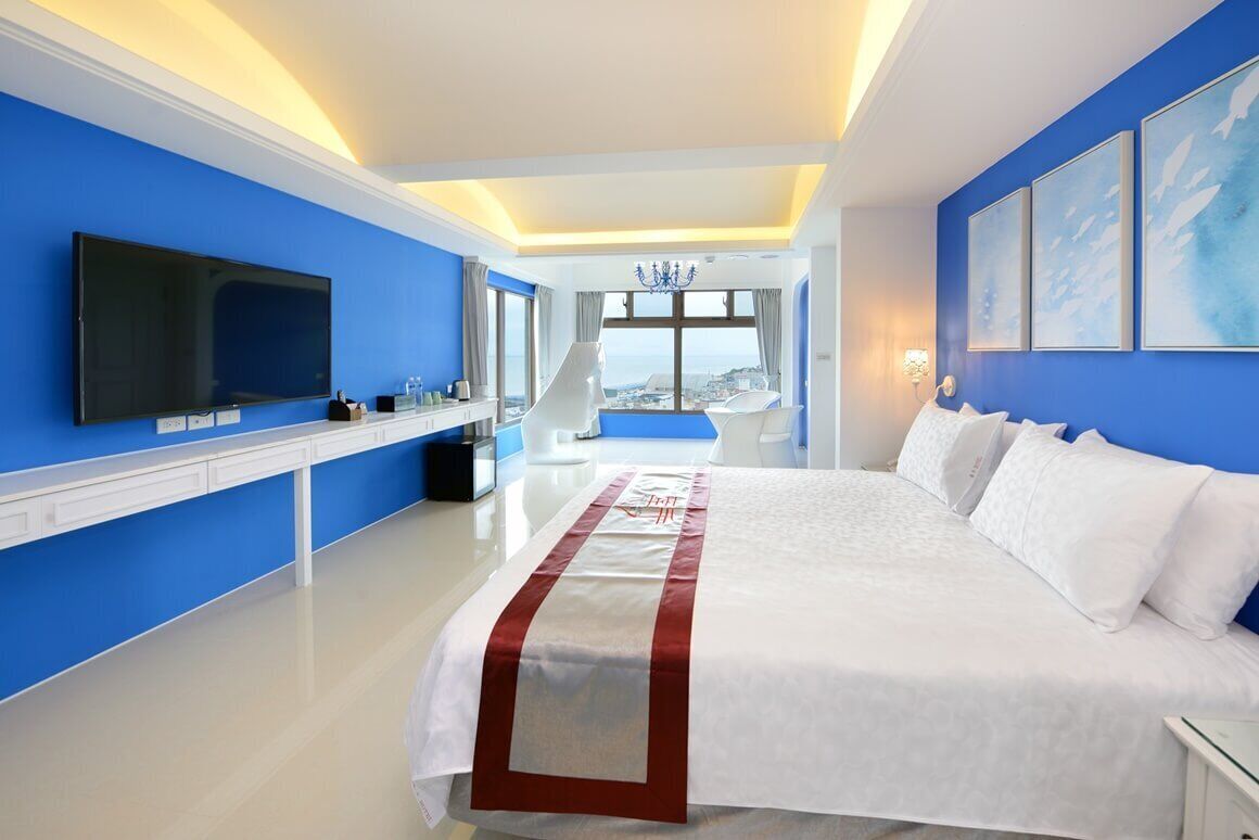 A modern hotel room with a large bed, a flat-screen TV, and a view of the city through large windows.