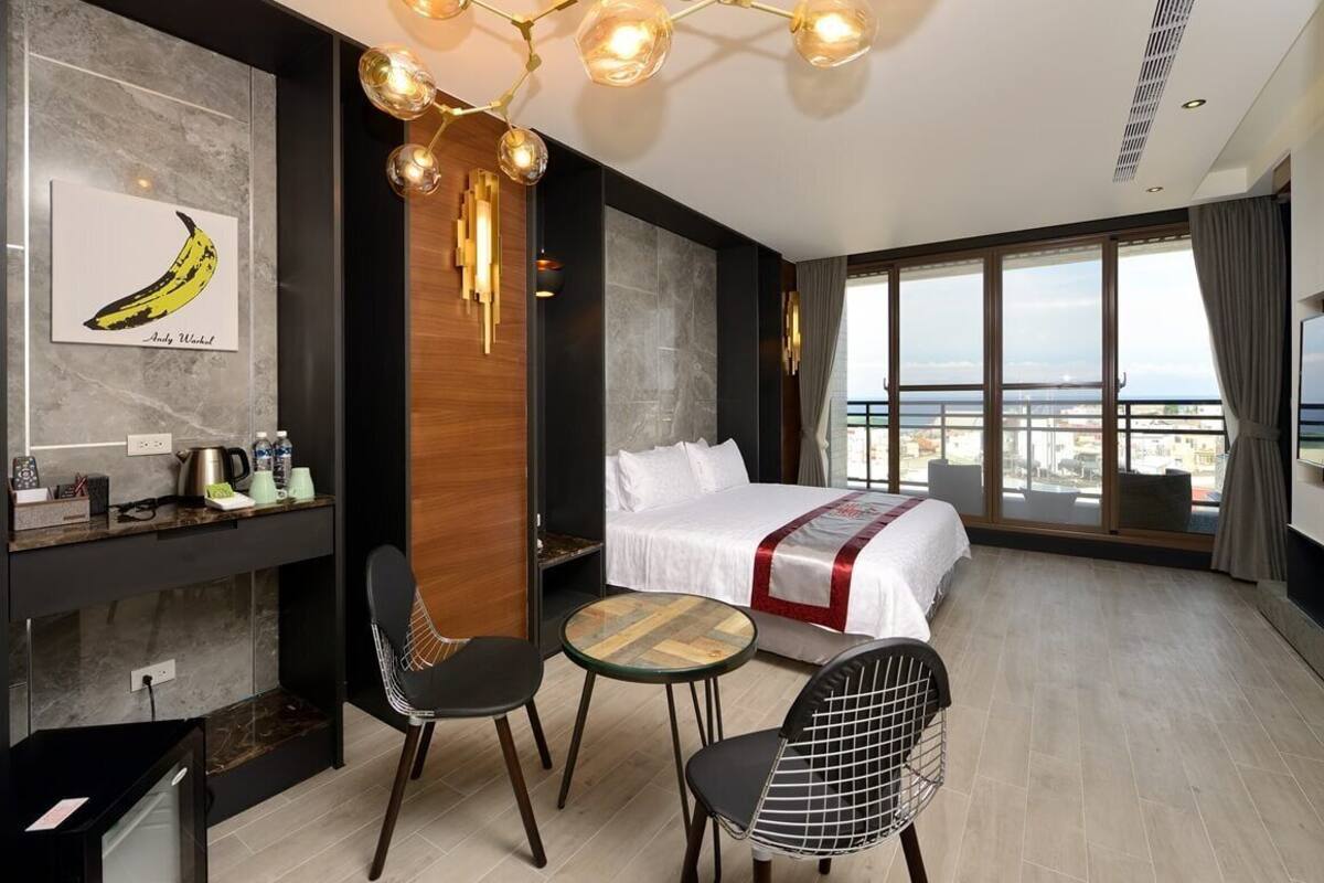 Una habitación de hotel moderna con una cama grande, una mesa de comedor, dos sillas, una pequeña cocina y vistas a la ciudad.