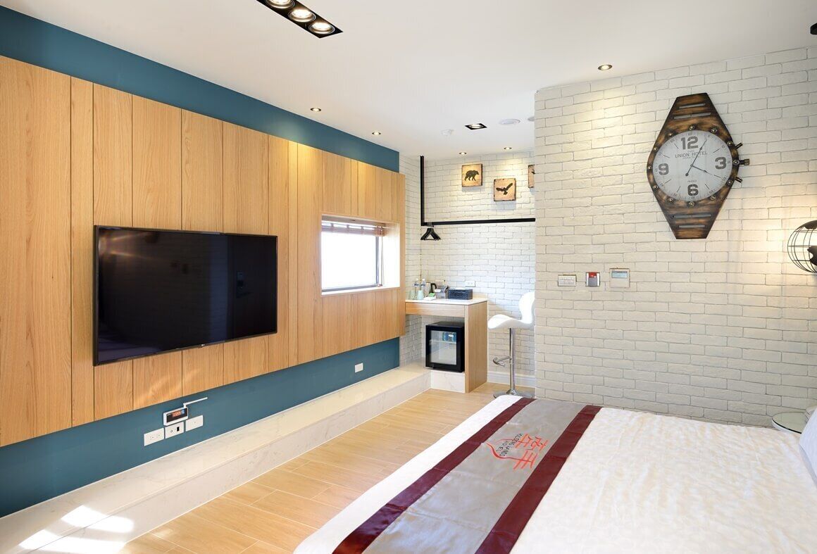 Una habitación de hotel moderna con un televisor de pantalla plana grande, un panel de pared de madera, una pared decorativa de ladrillo, un reloj original y una cama con colcha roja y blanca.
