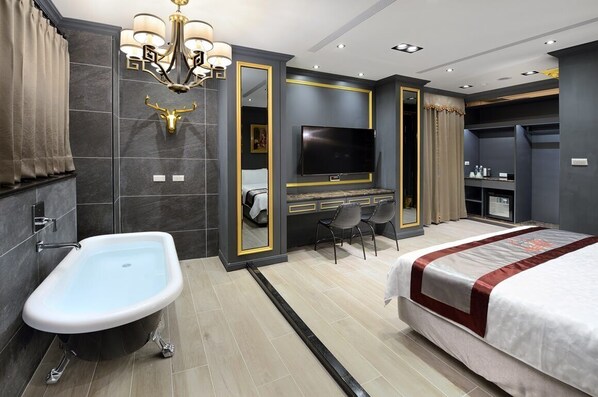 A modern hotel room with a bathtub, a large bed, a TV, and a desk with chairs.