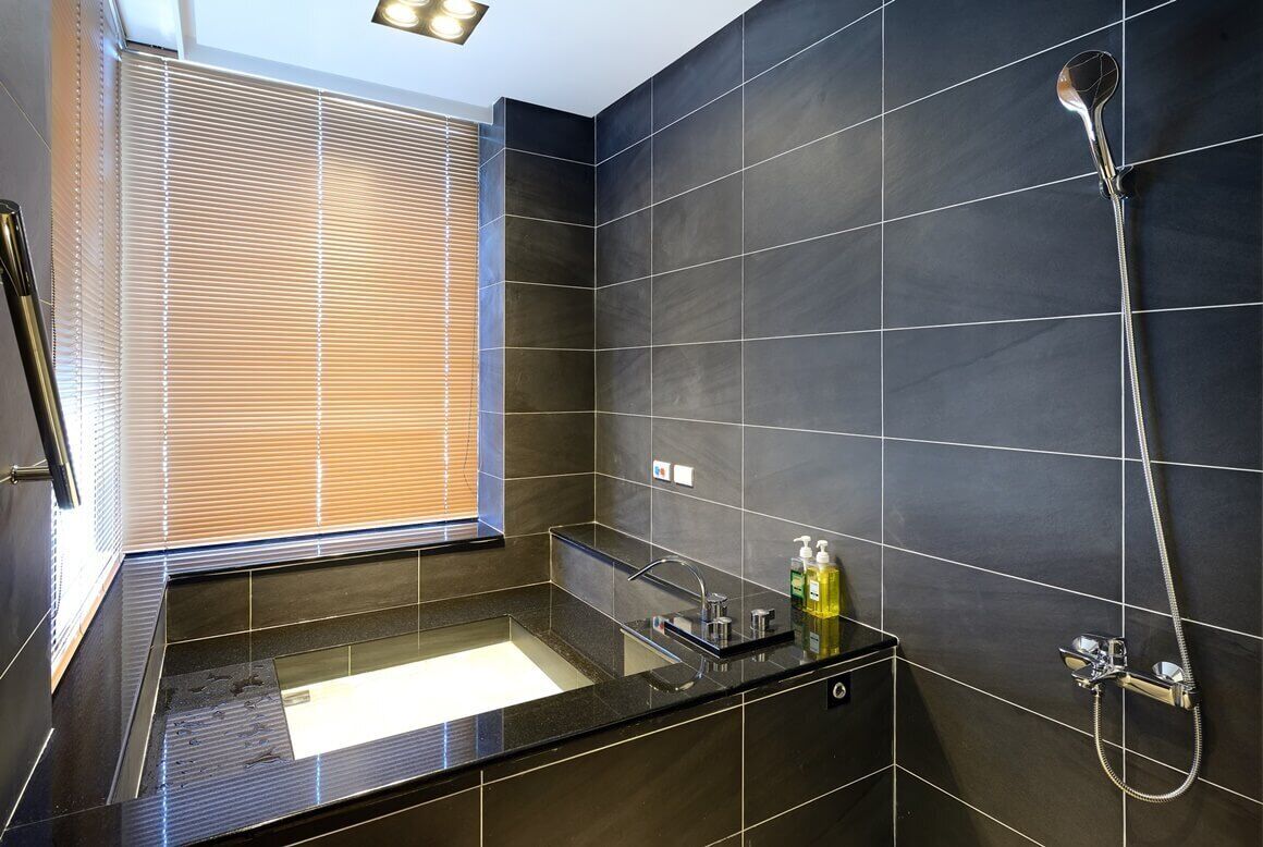 A modern bathroom with dark tiles, a glass shower enclosure, a sink with a mirror, and a window with blinds.