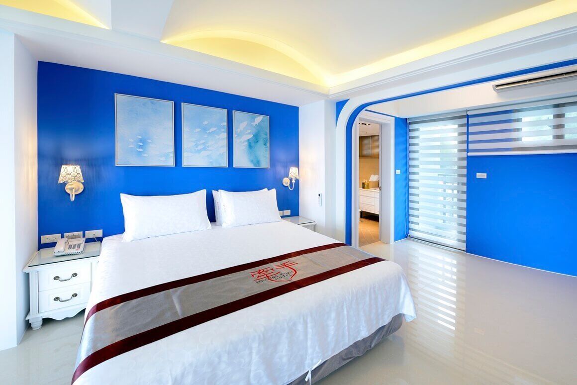 A modern hotel room with a large bed, a blue accent wall, and a view of the outside.