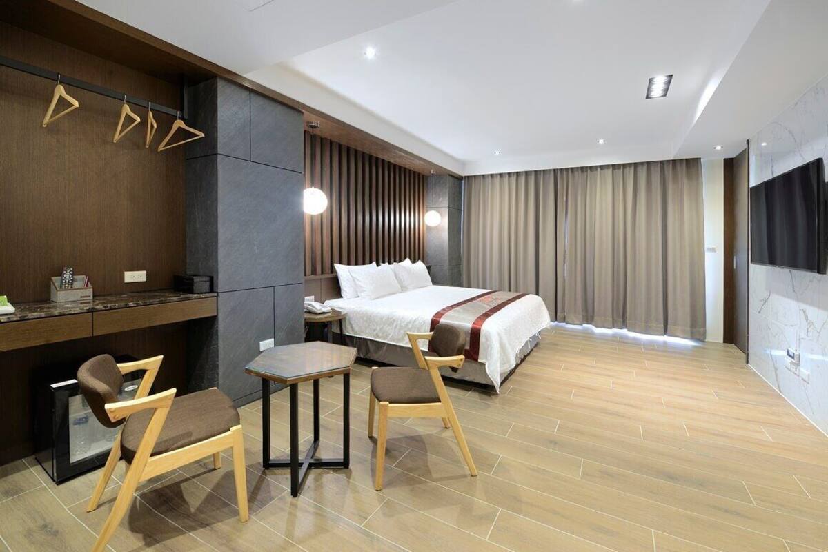 Una habitación de hotel moderna con cama, escritorio con sillas, televisor y un gran ventanal con cortinas.