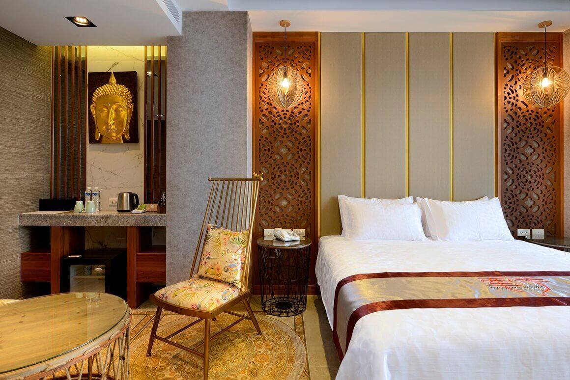 A hotel room with a bed, a chair, a small table, a desk, and a Buddha statue.