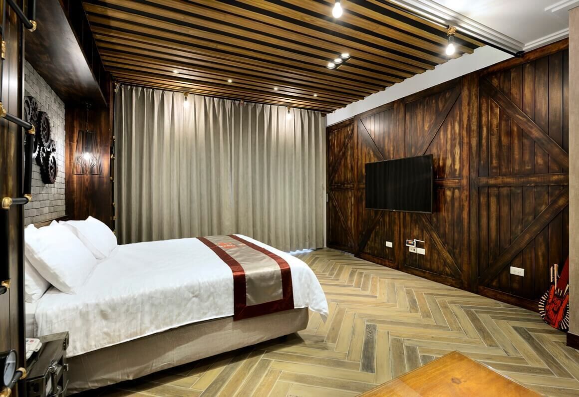A hotel room with a bed, wooden walls, a TV mounted on the wall, and a herringbone floor pattern.