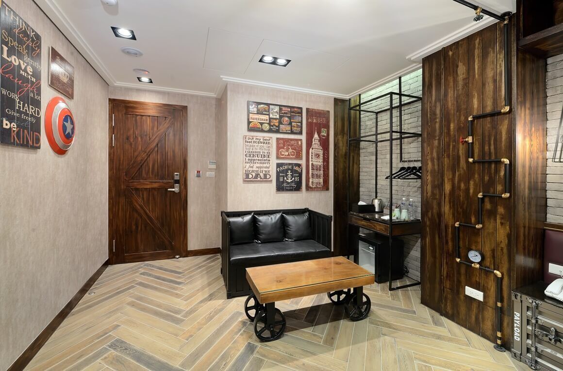 A hotel lobby with a wooden door, a black leather sofa, a wooden coffee table, and a wall with various posters.