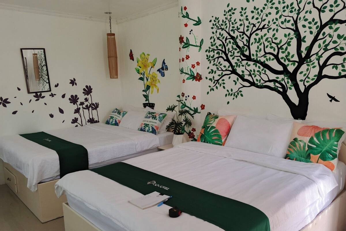 A hotel room with two beds, each with a green bedspread, white bedding, and a small table with a notebook and pen.