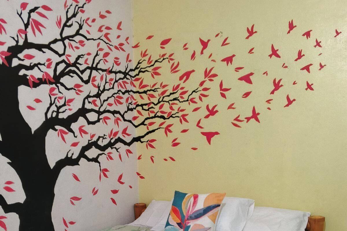 A bed with a floral bedspread, a colorful pillow, and a wall mural of a tree with red leaves.