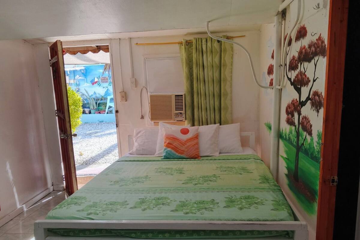 A bedroom with a bed, pillows, a window with curtains, a radiator, and a wall with a tree painting.