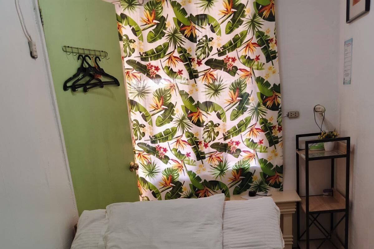 A small, neatly made bed with a floral-patterned bedspread and pillows, a green wall, a curtain with tropical leaf and flower prints, and a small shelf with a plant.