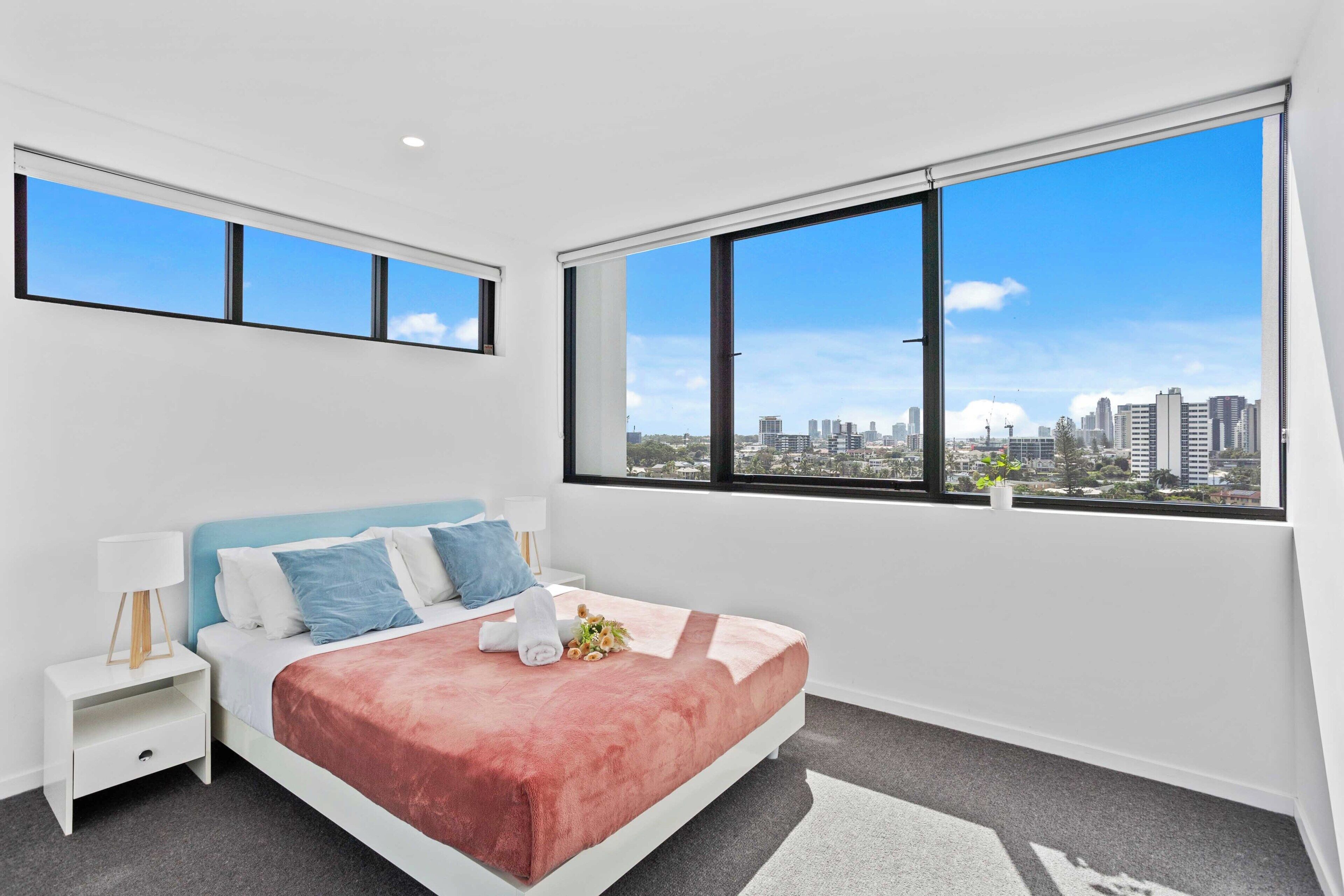 A modern bedroom with a city view, a bed with a pink blanket, a nightstand, and a lamp.