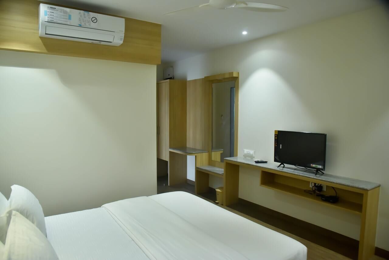 A hotel room with a bed, a TV on a wooden shelf, a mirror, and an air conditioning unit.