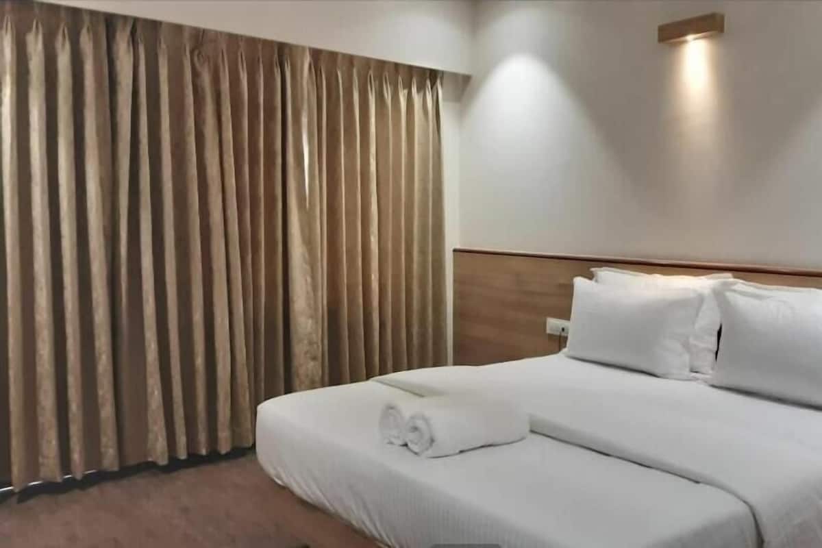 A hotel room with a neatly made bed, a large window with curtains, and a wall-mounted light fixture.