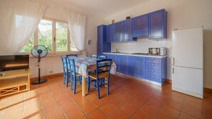A kitchen with blue cabinets, a tiled floor, a dining table with chairs, a fan, and a window with curtains.