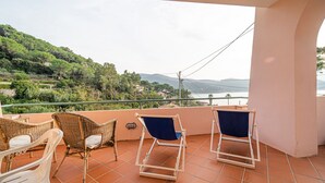 A balcony with wicker chairs and a terrace table, overlooking a scenic hillside and water body.