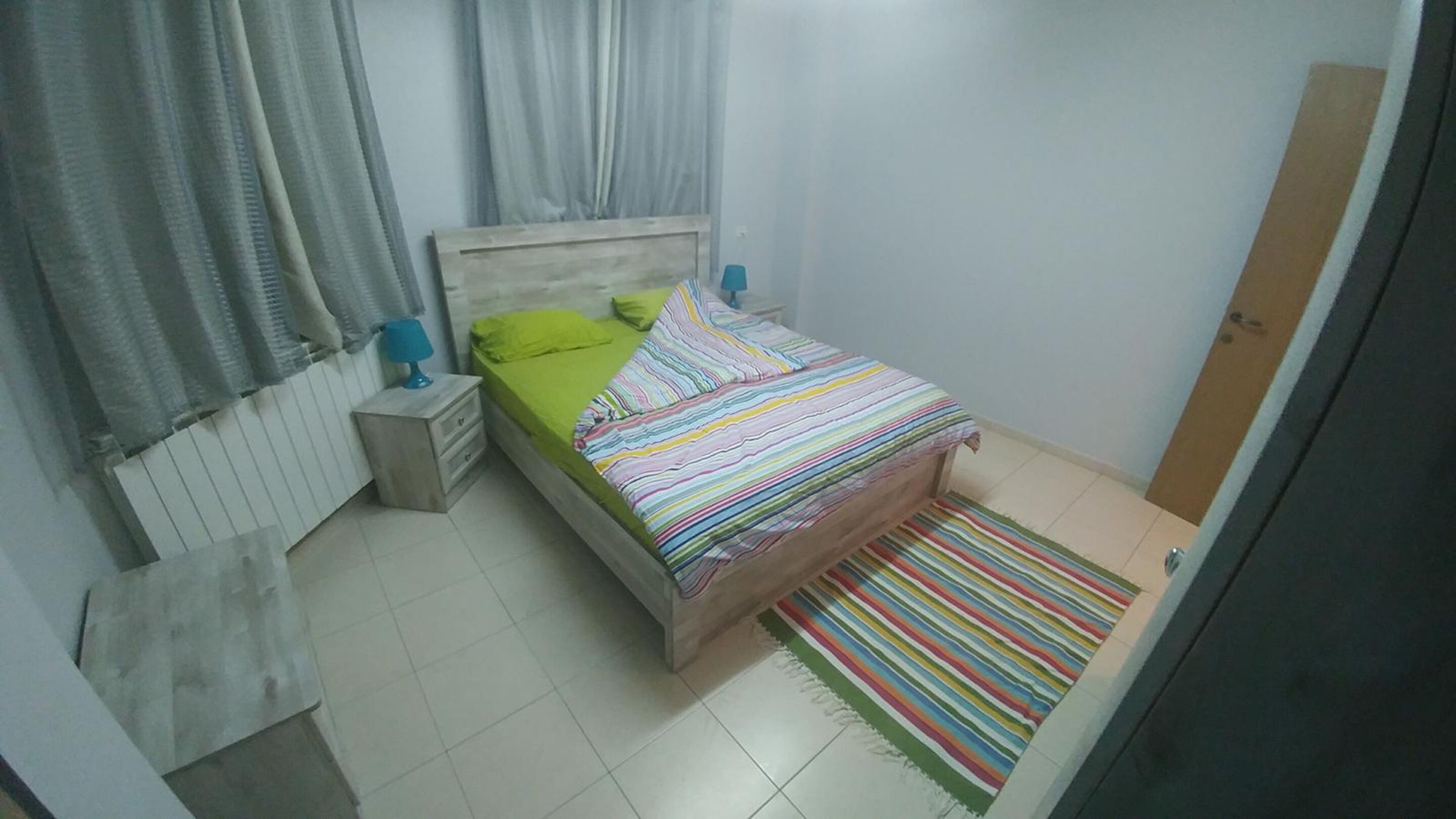 Ein kleines, ordentlich eingerichtetes Schlafzimmer mit einem Einzelbett, einem Nachttisch, einer Lampe und einem farbenfrohen Streifen Teppich.