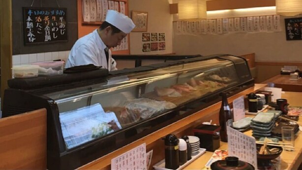 A sushi restaurant interior with a chef at the counter, a display case with seafood, and a menu board on the wall.