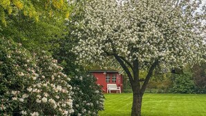 A red building with a white bench in front, surrounded by lush greenery and flowering trees.