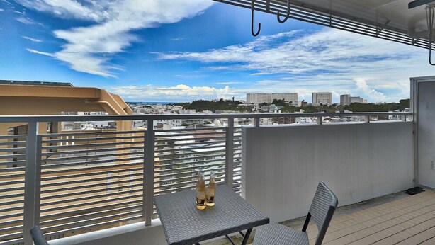 Un balcon avec une table et des chaises, donnant sur un paysage urbain avec des bâtiments et un ciel dégagé.
