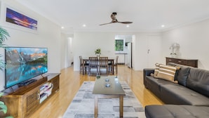 A modern living room with a flat-screen TV, wooden entertainment unit, a dining area with a table and chairs, a black leather sofa, and a ceiling fan.