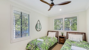 A bedroom with two single beds, a ceiling fan, a bedside table with a lamp, and a window with blinds.