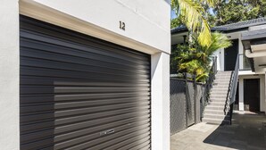 A modern building with a closed roller shutter garage door, a staircase leading to an upper level, and a fenced area with a palm tree.
