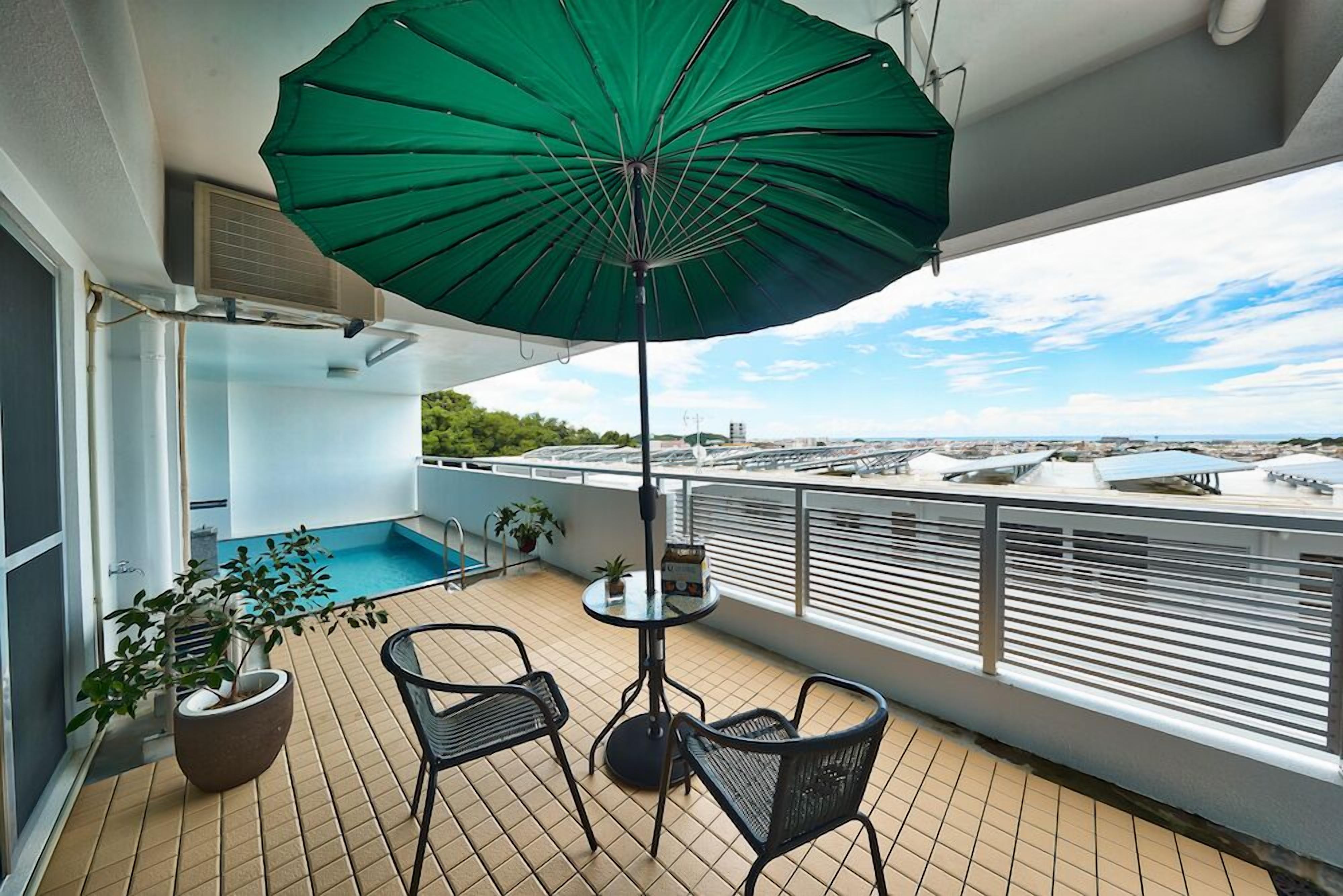 Varanda com guarda-sol verde, uma mesa pequena, duas cadeiras, um vaso de planta e uma piscina.