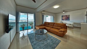 A spacious living room with a brown leather sofa, a round coffee table, and a large window offering a view of the outdoors. - FreedomHills A (Tomigusuku)