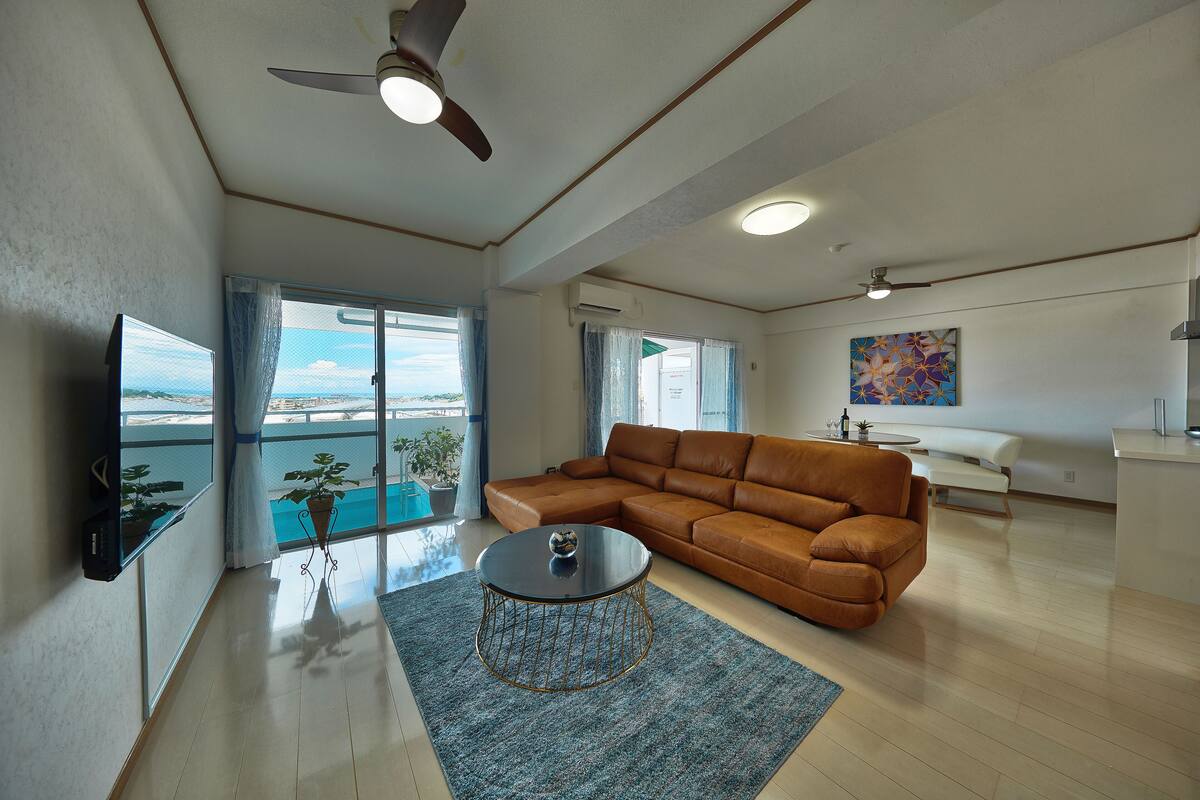A spacious living room with a brown leather sofa, a round coffee table, and a large window offering a view of the outdoors.