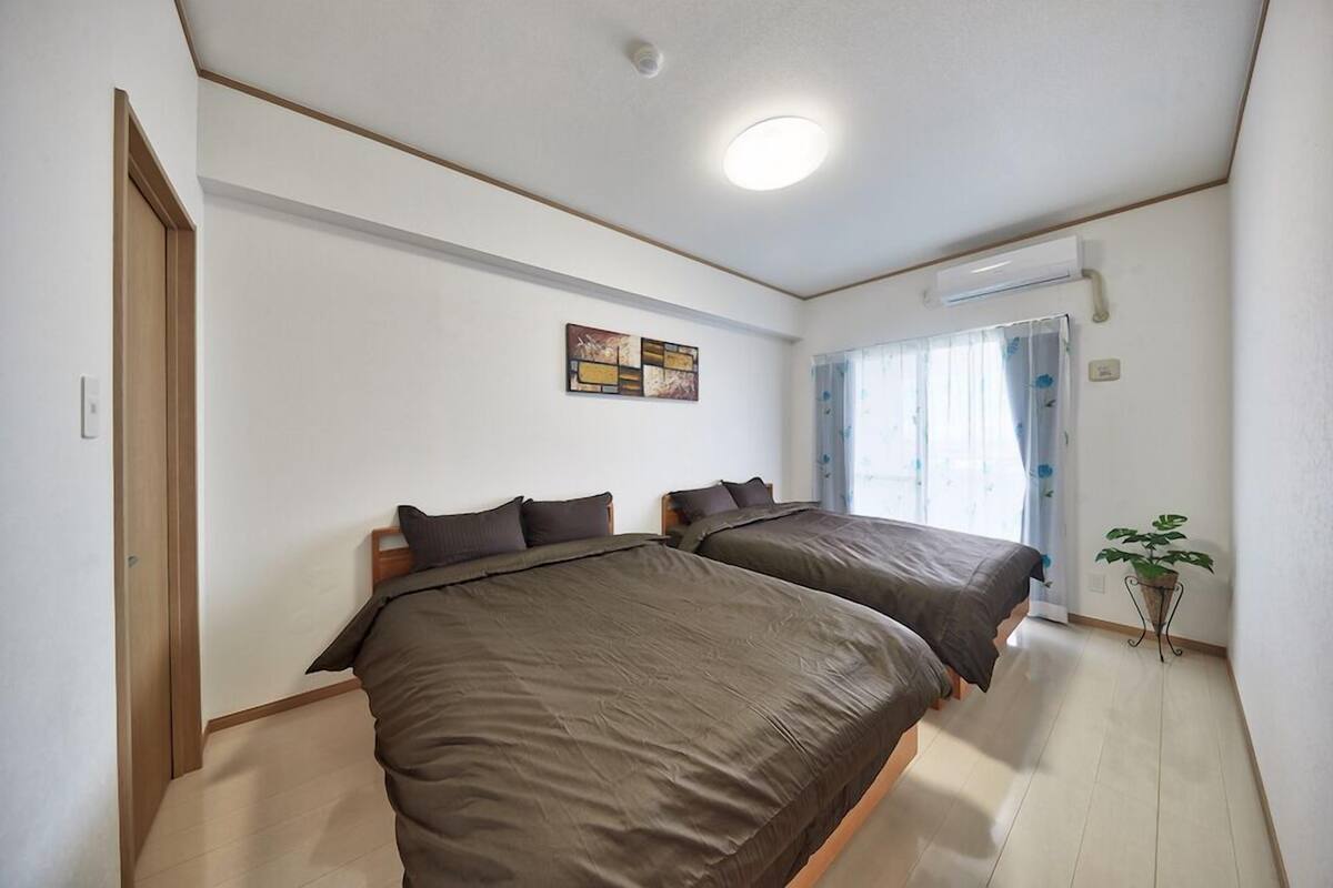 A bedroom with two beds, a ceiling light, a window with curtains, and a small potted plant.