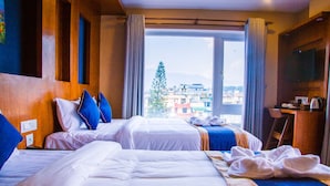 A hotel room with two beds, a large window with a view of buildings and a tree, and a TV on the wall. - Himalayan Suite Hotel (Kathmandu)