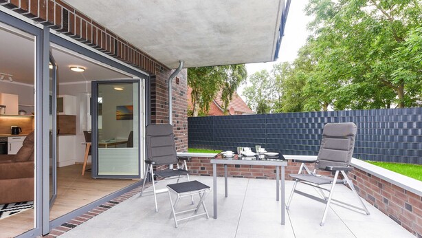 An outdoor patio with a table and chairs, a sliding glass door leading to a kitchen, and a brick wall with a fence. - Vacation apartment 14 - Haus Seeperle (Esens)