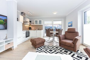 Una sala moderna con un sillón de cuero marrón, una mesa de comedor con sillas, una zona de cocina con armarios blancos y un ventanal que proporciona luz natural.
