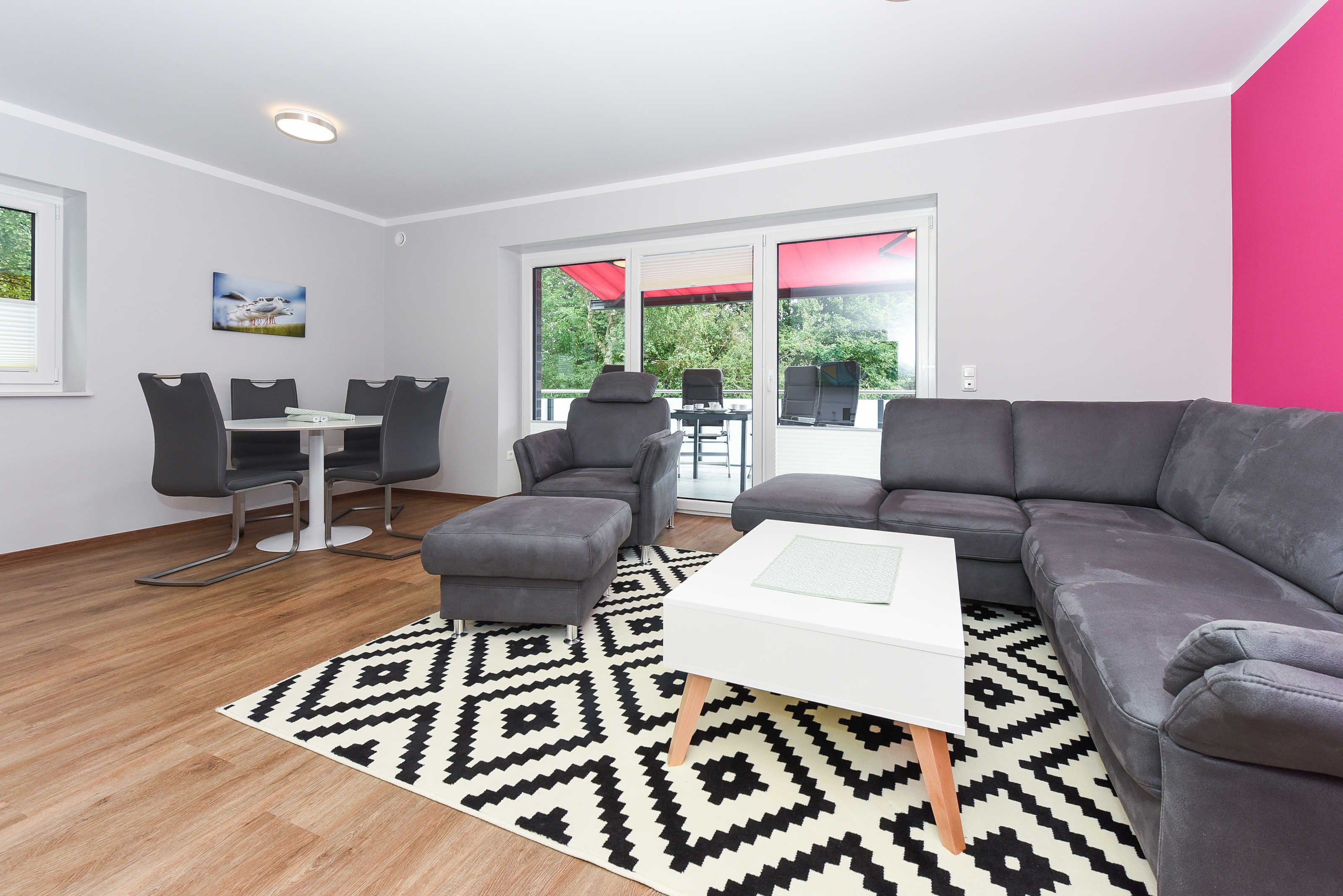 Een moderne woonkamer met een grijze bank, een witte salontafel en een vloerkleed met een patroon. Er is een eethoek met een tafel en stoelen, en een groot raam met een rode paraplu.