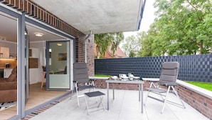 An outdoor patio with a table and chairs, a sliding glass door leading to a kitchen, and a brick wall with a fence. - Vacation apartment 18 - Haus Seeperle (Esens)