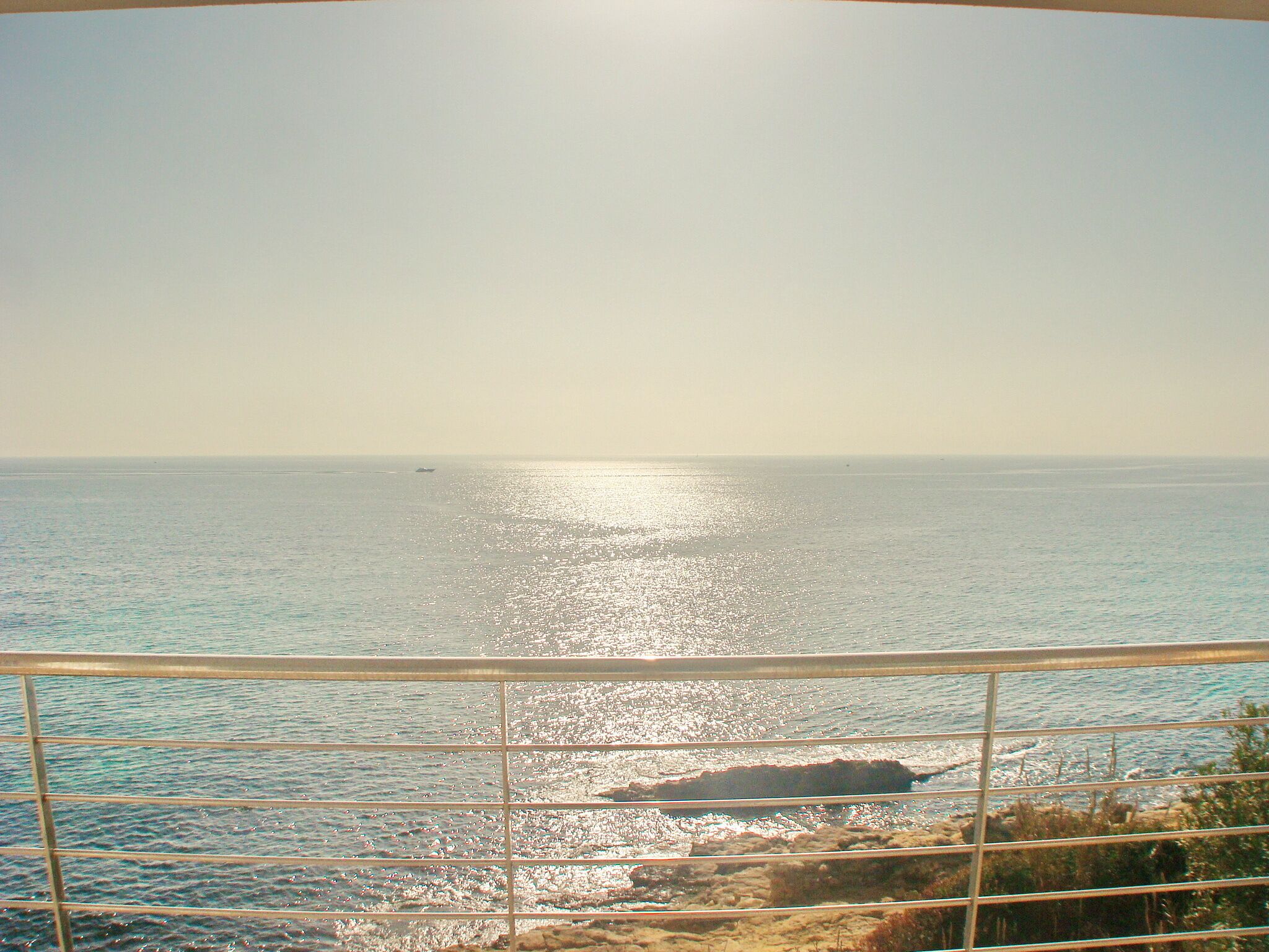A coastal view with a metal railing in the foreground, a clear sky, and calm sea.