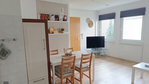 A modern kitchen with white cabinets, a dining table with two chairs, a TV on a glass stand, and a window with blinds. - Beautiful bungalow near the Baltic Sea, family-friendly and quiet (Nienhagen)