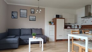 A modern living room with a grey sofa, a white coffee table, and a dining area with a wooden table and chairs. There is a kitchen with white cabinets and a microwave on top. - Beautiful bungalow near the Baltic Sea, family-friendly and quiet (Nienhagen)