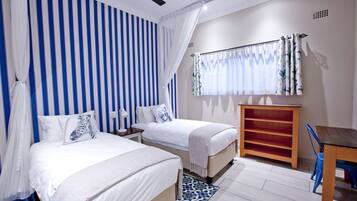 Un dormitorio con dos camas, un ventilador de techo, una ventana con cortinas, una mesita de noche y un pequeño armario de madera.