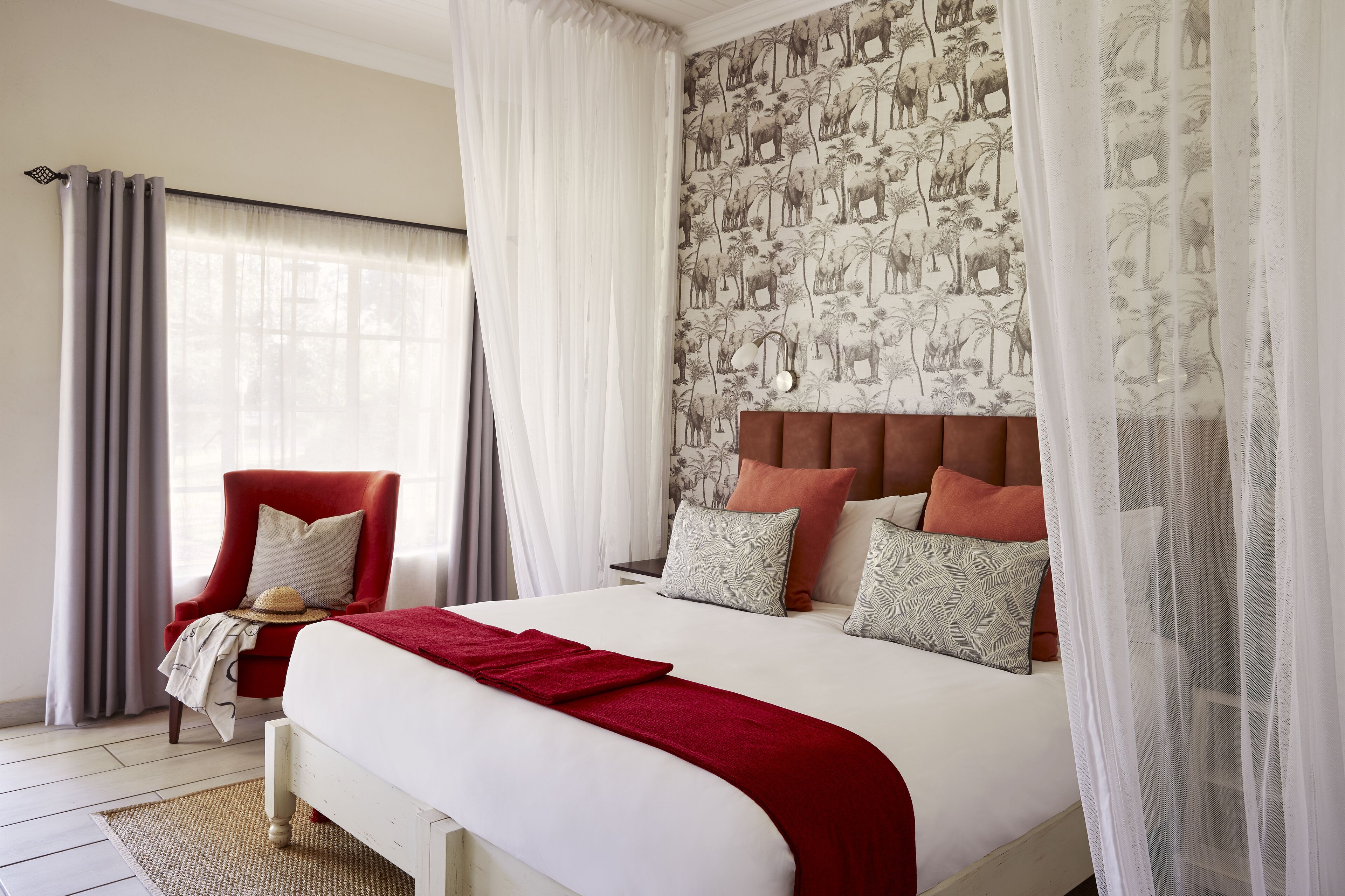 A neatly made bed with a red throw blanket, a red armchair with a straw hat, and a patterned wallpaper.
