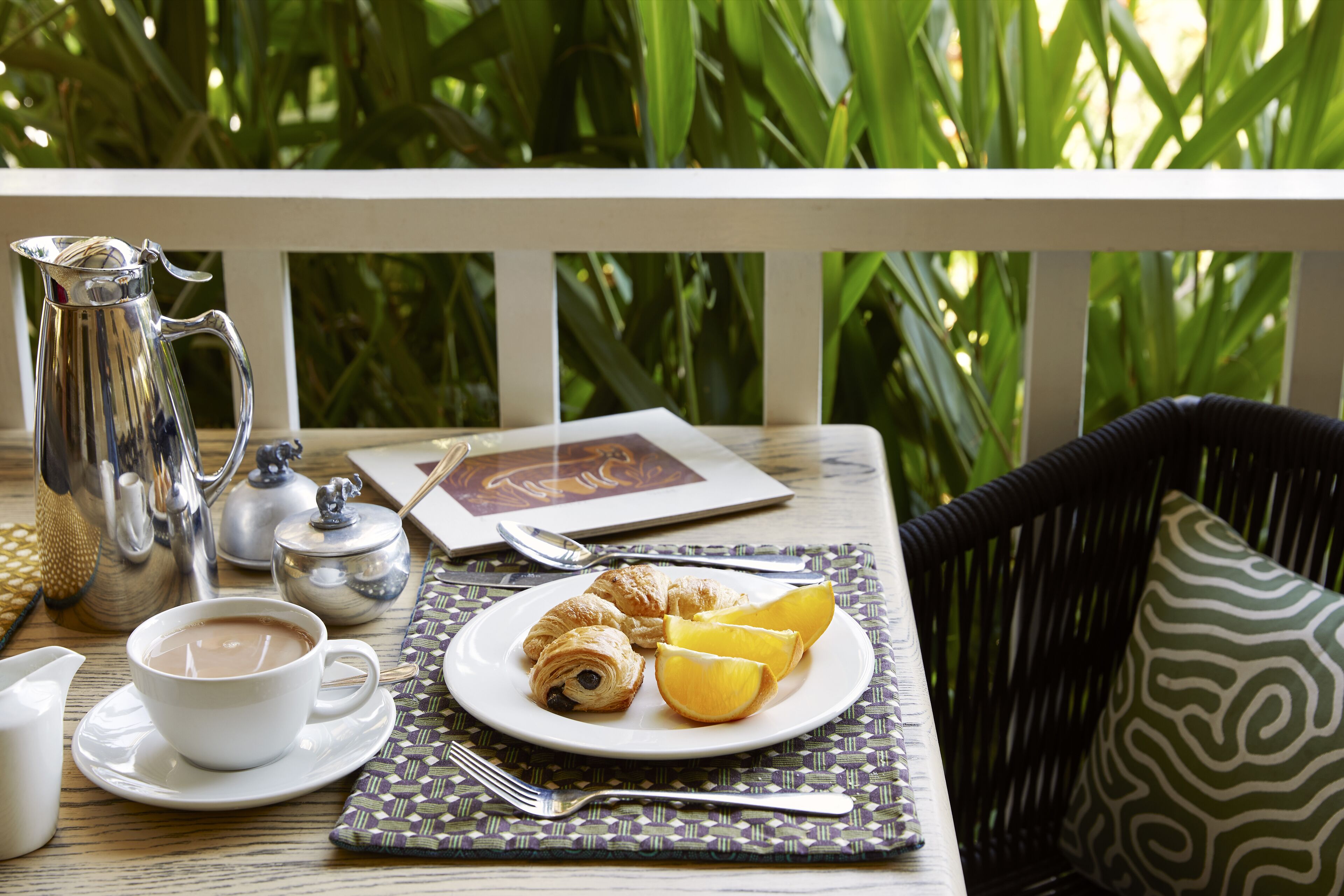 A table set with a cup of coffee, croissants, and orange slices on a placemat, with a silver pitcher and a teapot on the table.