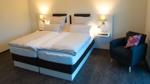 A double bed with white bedding, two bedside tables with lamps, a dark armchair with a patterned pillow, and a wooden floor. - Hof Hahnenberg (Wagenfeld)