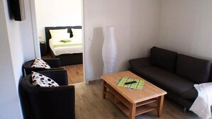 A small living area with a dark sofa, a wooden coffee table, and a chair with a polka dot cushion. A bed with white bedding is visible through an open door in the background. - Hof Hahnenberg (Wagenfeld)