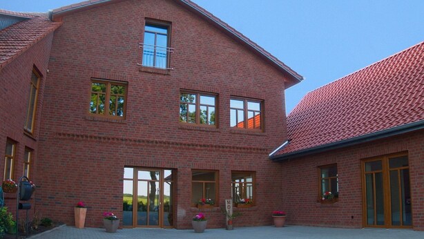 A two-story brick building with large windows, a covered patio, and potted plants. - Hof Hahnenberg (Wagenfeld)