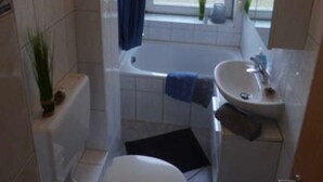 Ein kleines Badezimmer mit Toilette, Waschbecken und Badewanne. Ein Fenster mit Vorhang und ein Topf auf dem Fensterbrett.